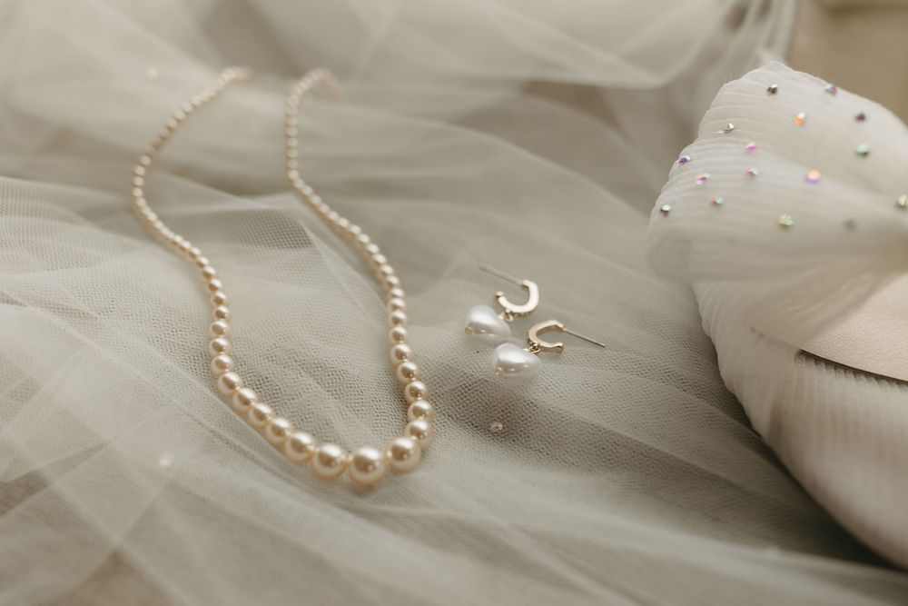 A delicate pearl necklace and crescent moon earring rest on soft tulle fabric beside white satin material.