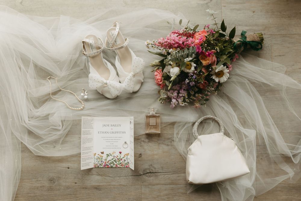 Wedding accessories including tulle veil, floral bouquet with pink blooms, invitation card, perfume bottle and white purse on wooden floor.