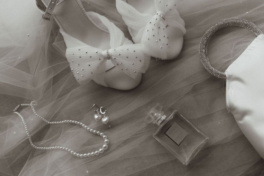 Wedding accessories including white bow shoes, pearl necklace and perfume bottle arranged on tulle fabric.