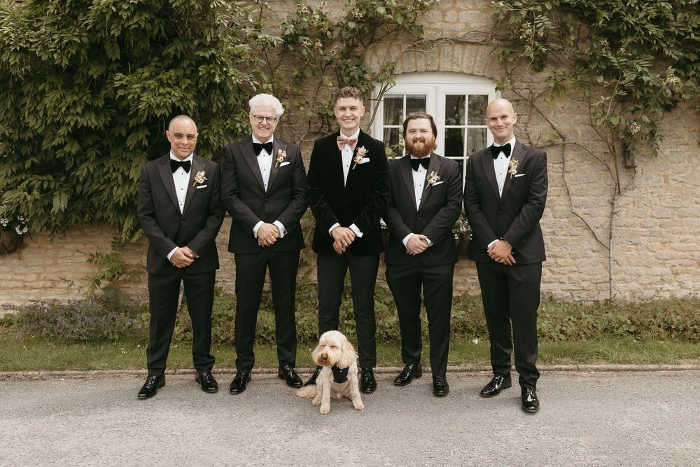 Groomsmen in black tuxedos standing with a small dog in formal attire at a brick venue.