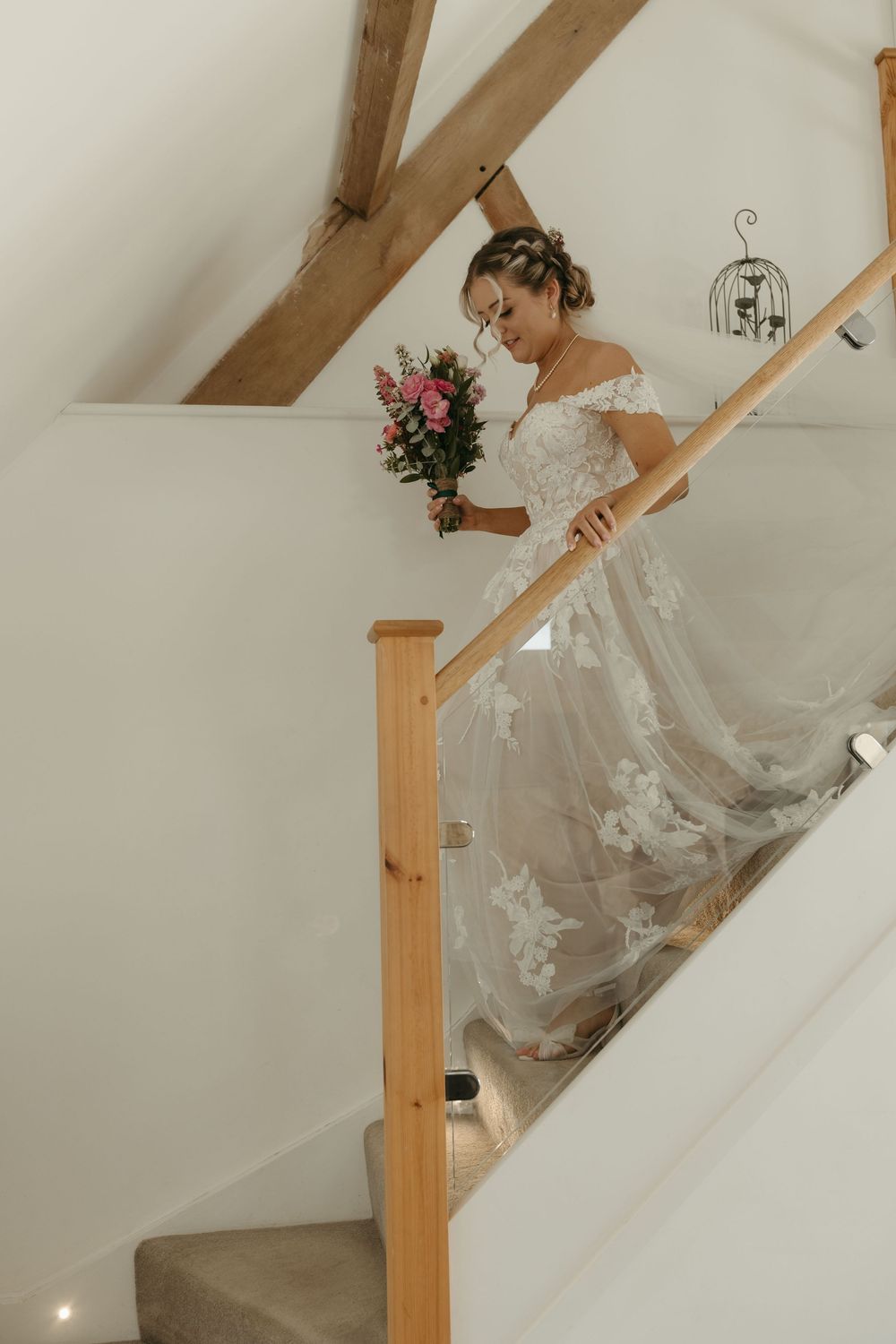 A bride in a lace wedding gown descends a staircase while holding a pink and white bouquet.