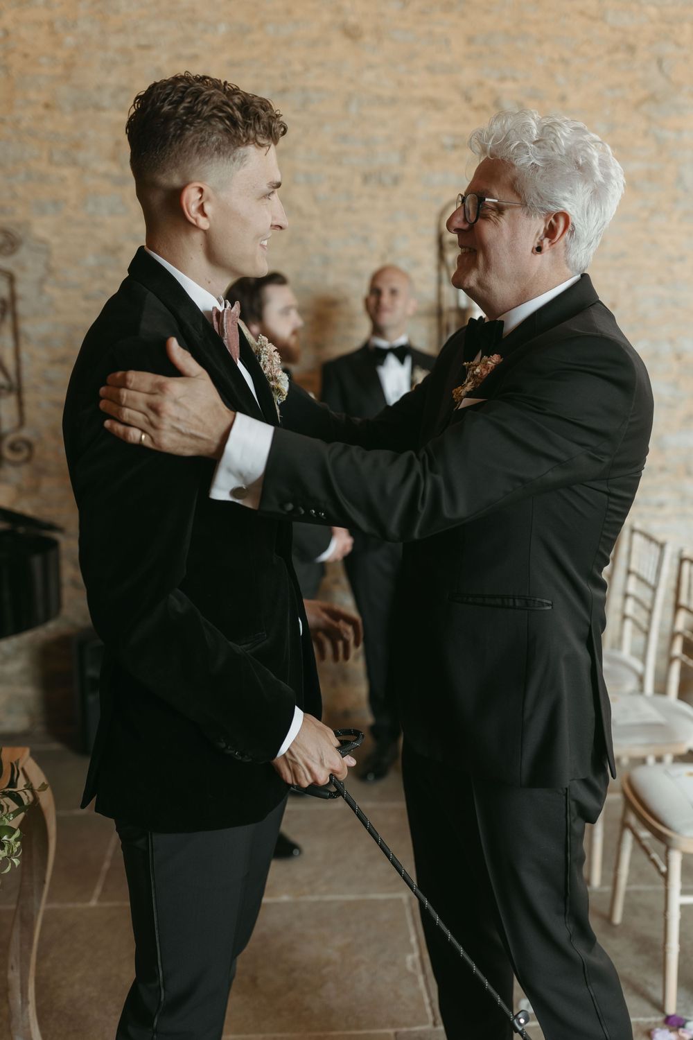 Two men in formal black suits share an emotional moment before a wedding ceremony in a brick-walled venue.