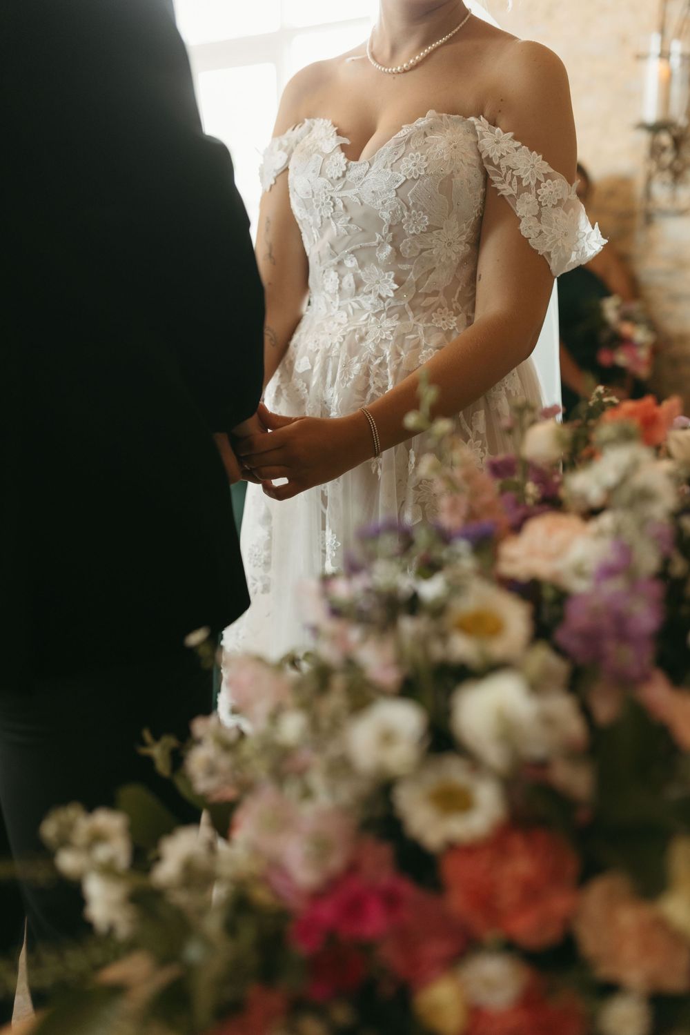 Detail shot of bride in lace off-shoulder wedding dress with colorful floral arrangements.
