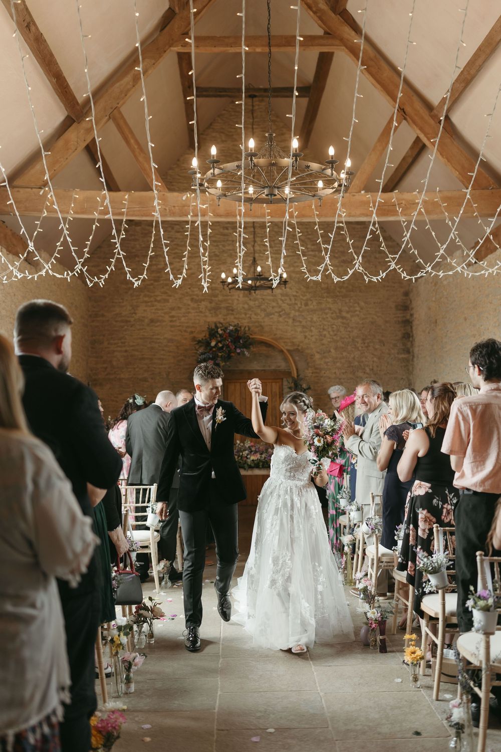 A festive barn wedding ceremony with twinkling string lights and exposed wooden beams overhead.