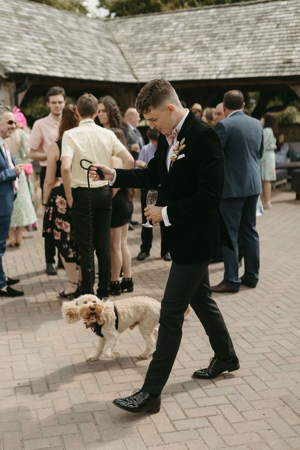 Wedding guest in formal attire walking a small dog on a leash at an outdoor reception.
