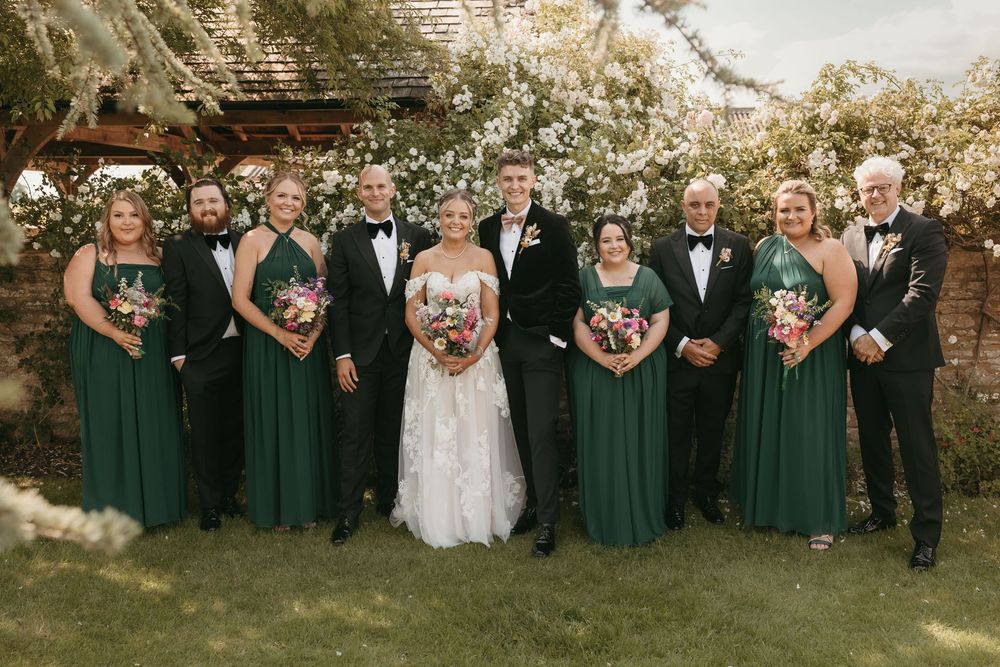 Wedding party poses outdoors in formal attire with bridesmaids in emerald green dresses and groomsmen in black suits.