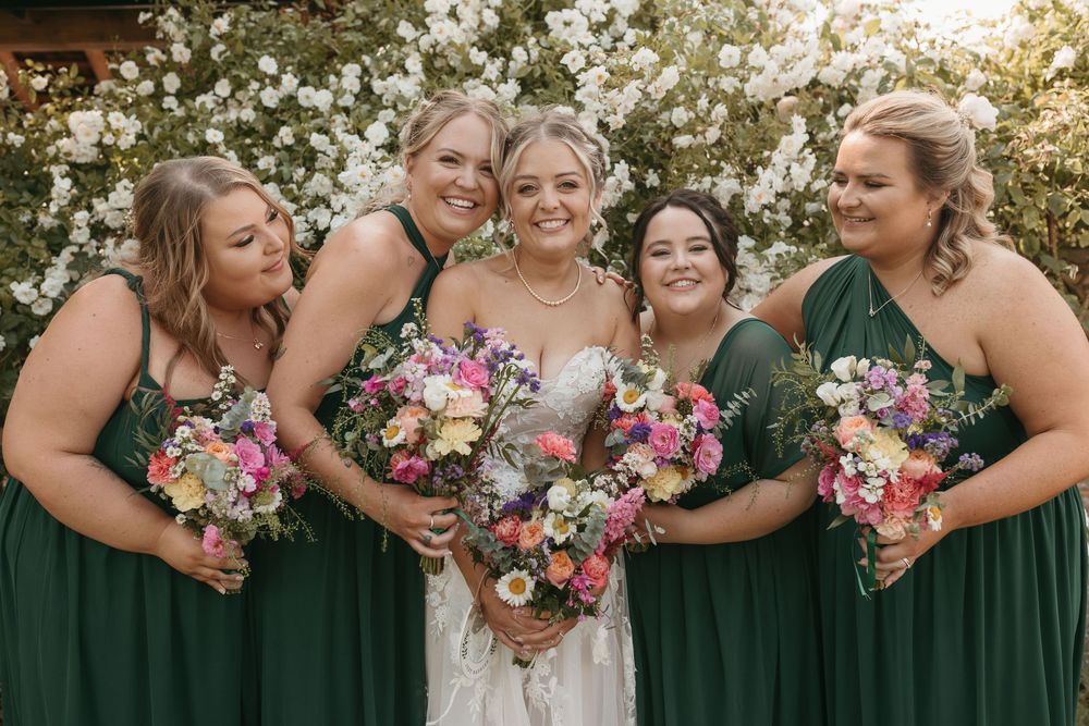 Bridesmaids in emerald green dresses holding pink and white bouquets at an outdoor wedding.