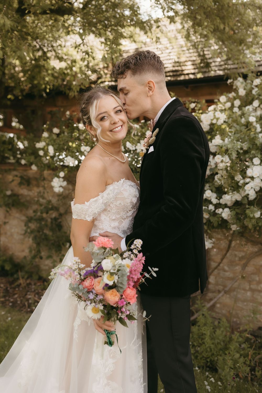 A romantic outdoor wedding portrait with pink and coral flower bouquet against a rustic stone wall backdrop.