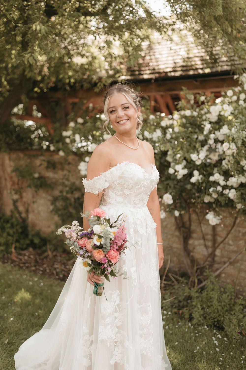 A beautiful off-shoulder lace wedding gown with a flowing train and pink floral bouquet in a garden setting.