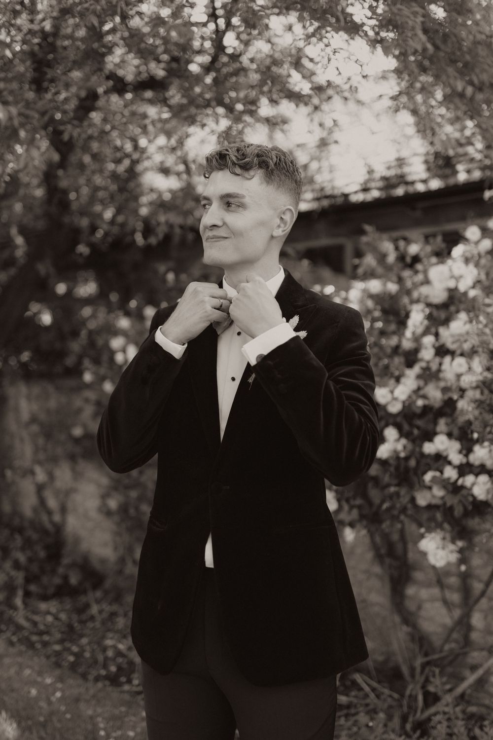 Black and white portrait of a well-dressed person in a velvet tuxedo adjusting their bowtie among blooming trees.