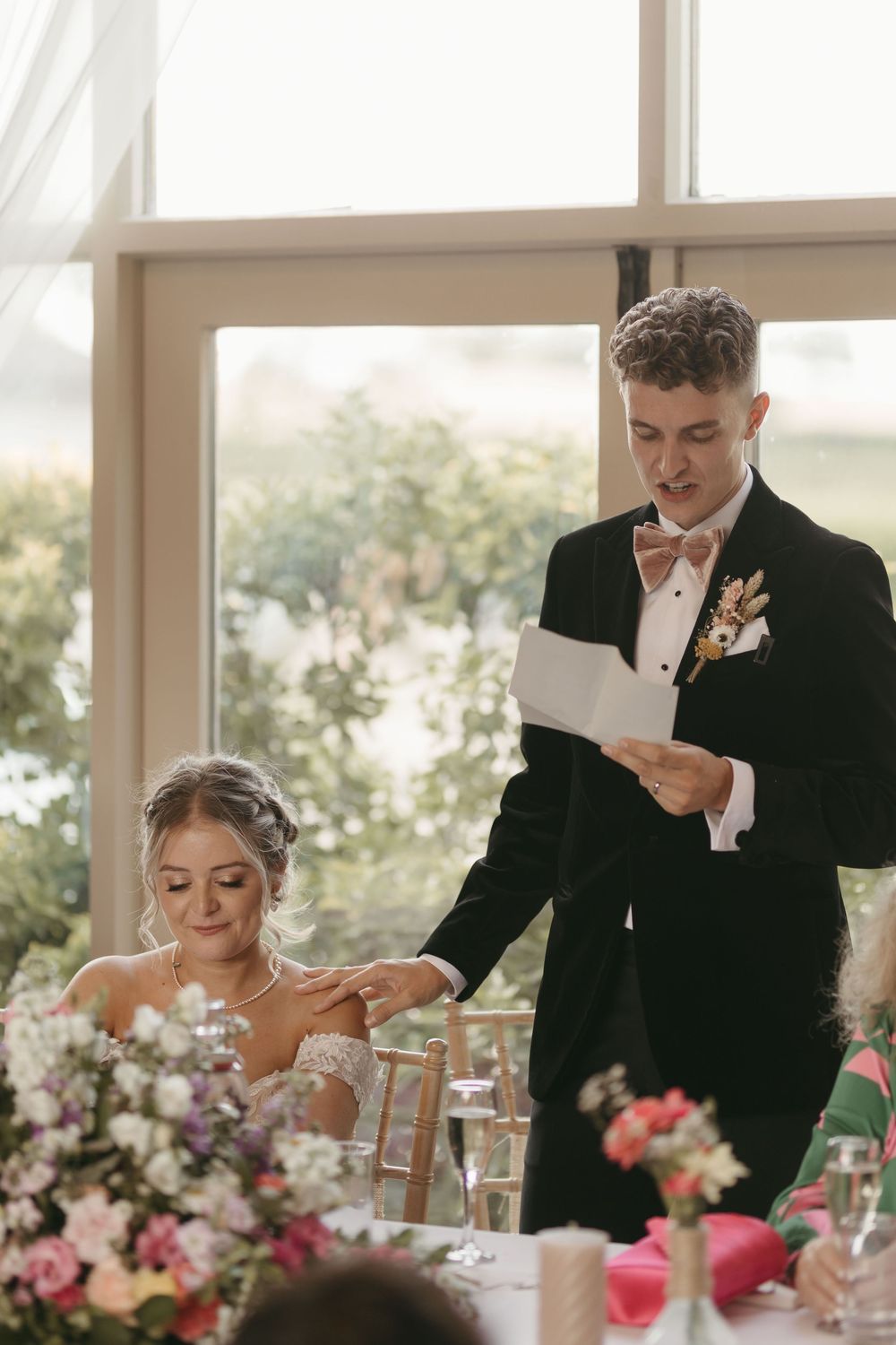 A reception scene with wedding toasts being given at an elegantly decorated table with pink floral centerpieces.