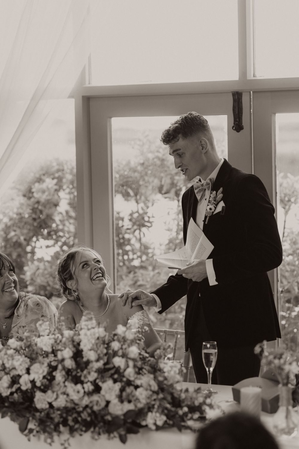 Black and white photo of a wedding celebration as laughter fills the air during speeches at a floral-decorated reception table.