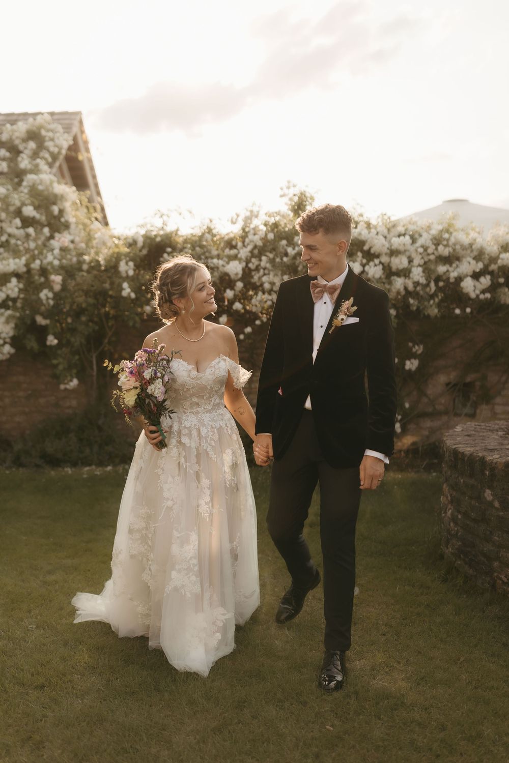 A couple in wedding attire walking together in a garden during golden hour with stone walls in the background.