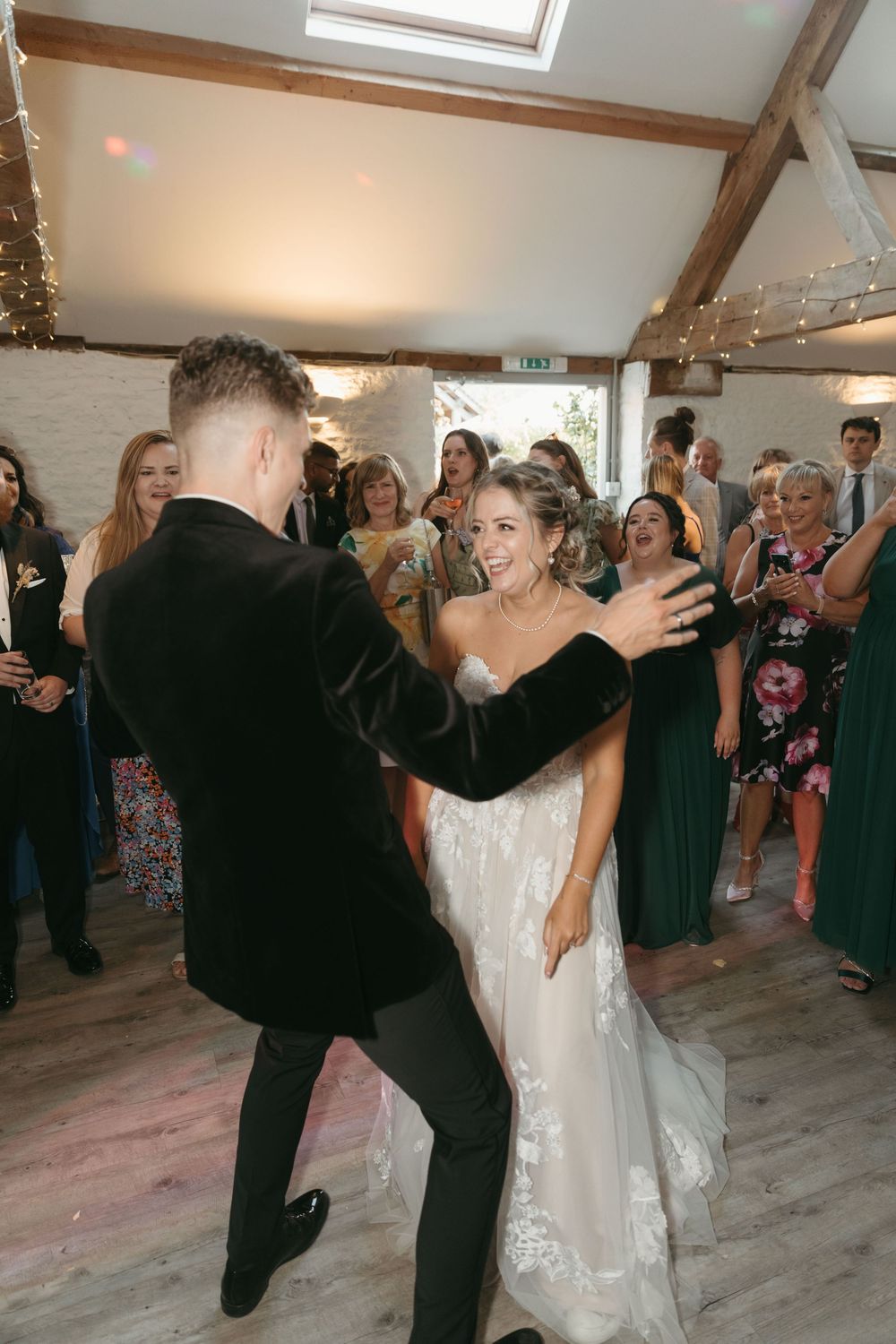 A joyful first dance moment between newlyweds in a rustic venue decorated with string lights as guests look on.