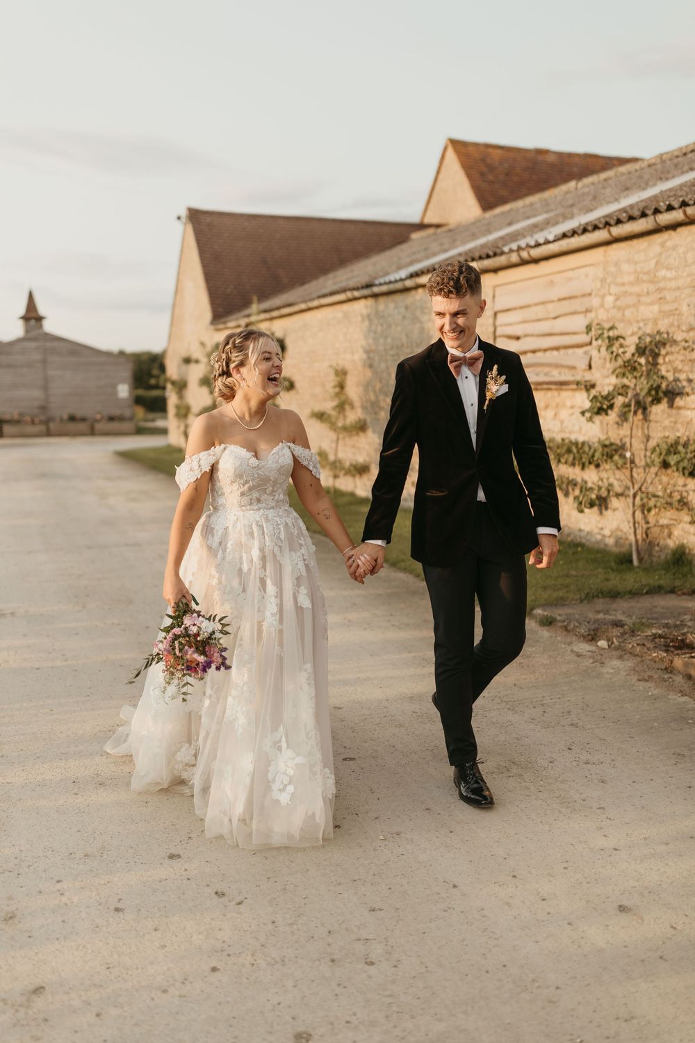 Couple walks hand in hand on dirt path by rustic buildings, bride in off-shoulder lace gown carrying floral bouquet.