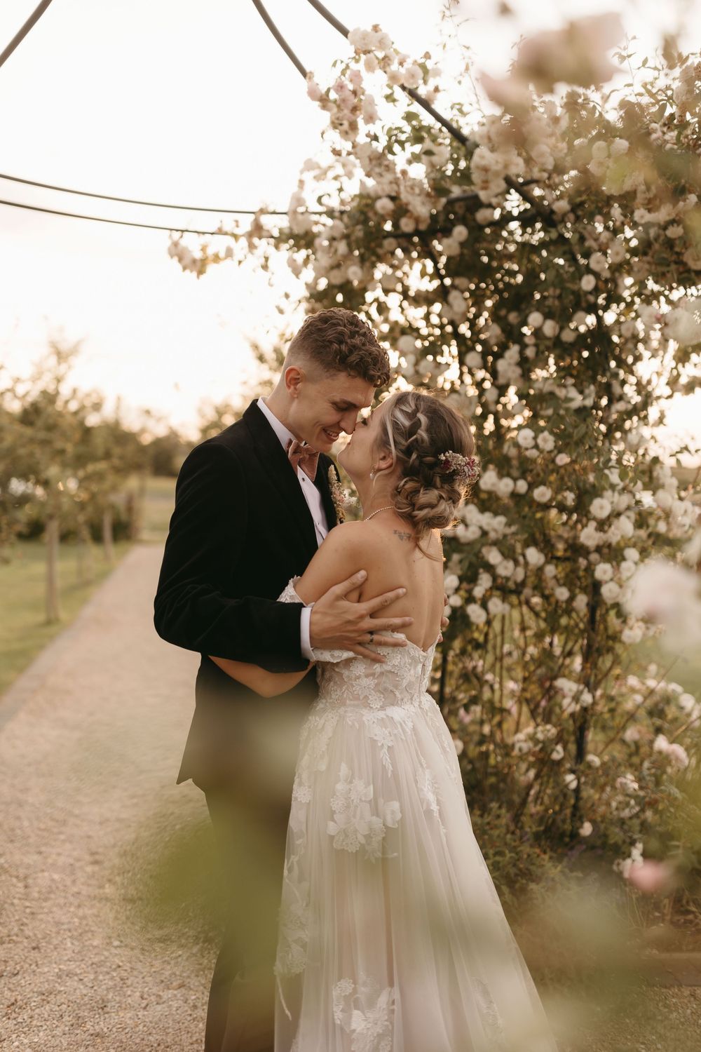 A romantic wedding couple embraces on a gravel path with blooming flowers and greenery at sunset.
