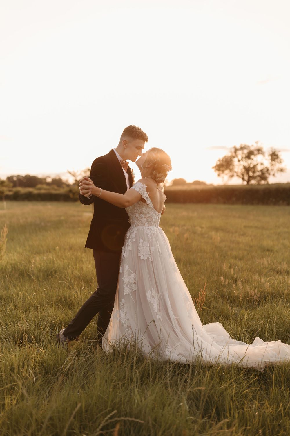 A romantic wedding couple dances together in a golden sunset-lit field with the bride wearing a flowing lace gown.