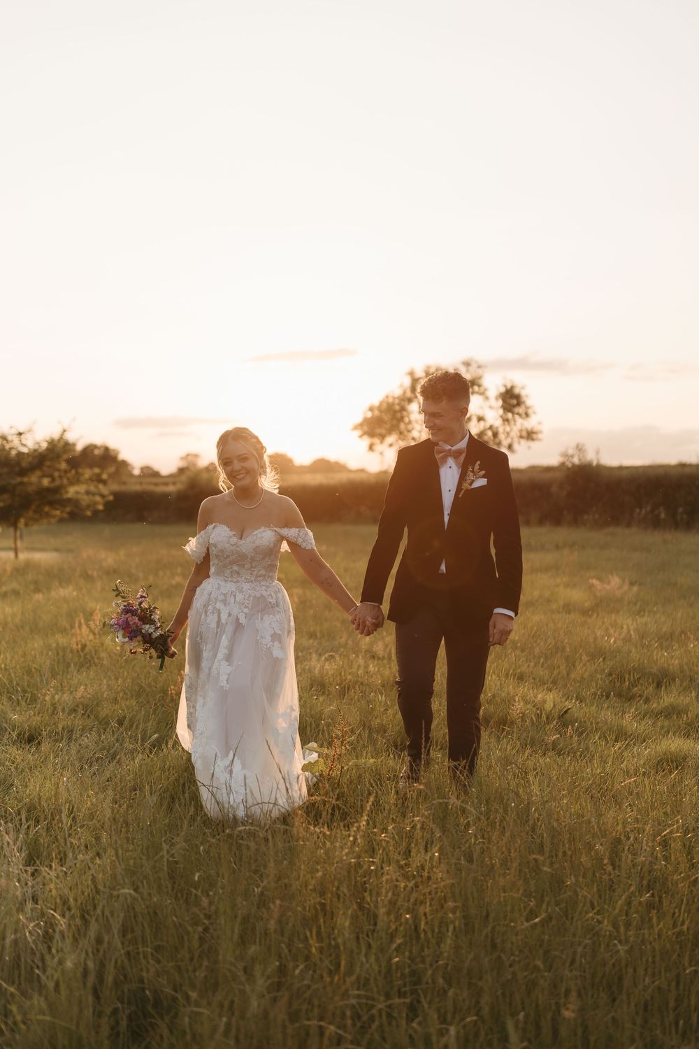 Newlyweds walking hand in hand through a golden sunlit meadow at sunset during their wedding photoshoot.