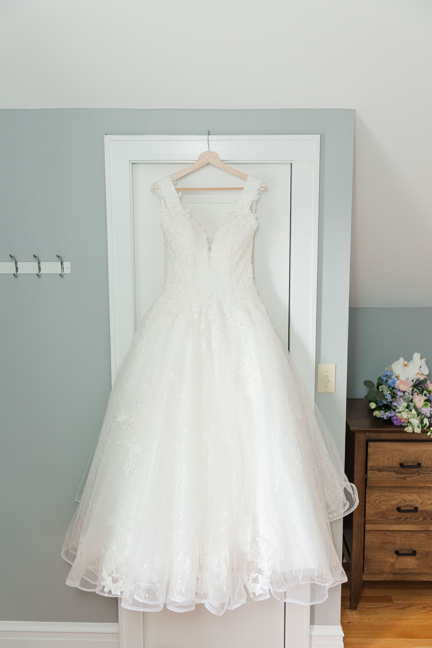 Elegant white wedding gown with full tulle skirt and lace details hangs displayed on wooden hanger against pale mint wall.