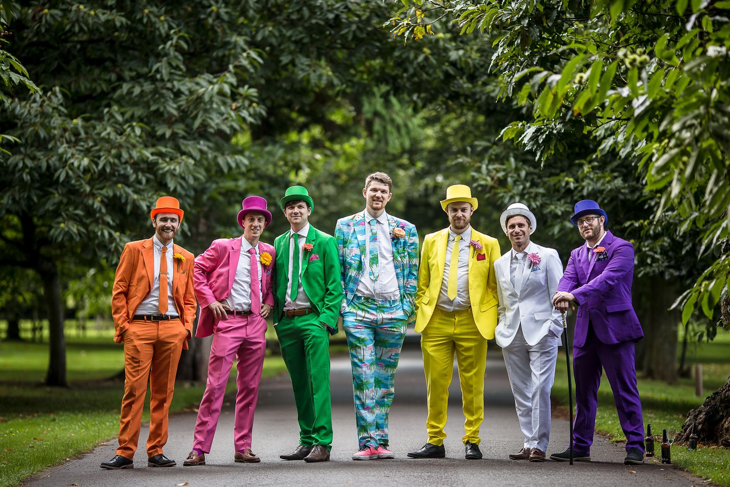 Groom and Groomsmen in vibrant colored suits stand together in a line outdoors against a green garden backdrop.