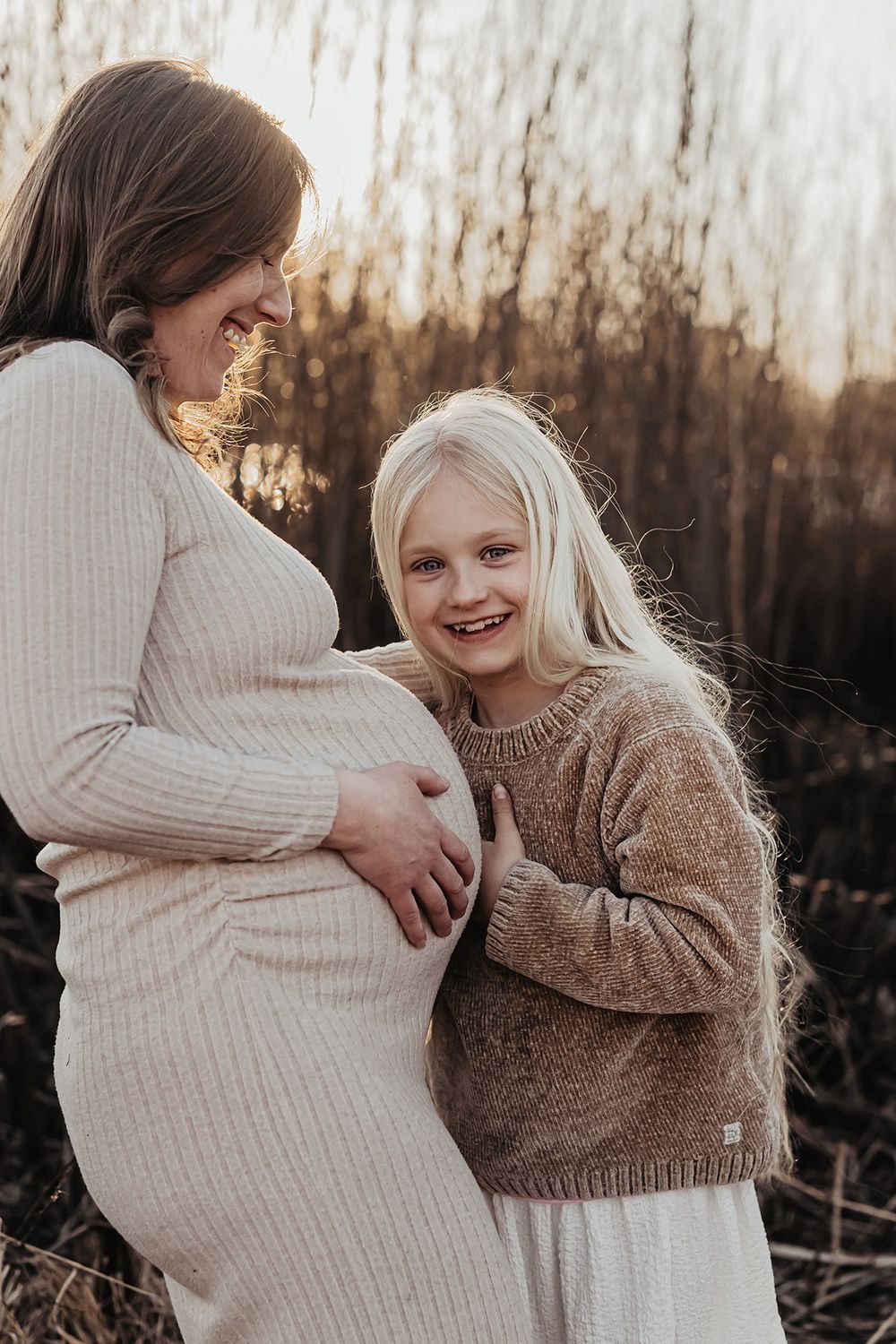 Gravidfoto med en mamma och hennes dotter, dottern hålla om magen och skrattar. Det är vackert motljus vid stranden, foto Lets Bloom Media.