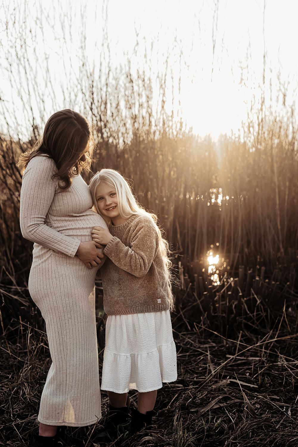 Gravidfoto i solnedgången vid vattnet på mamma och dotter som står nära varandra. Ljuset faller över dem och får bilden att glöda av värme.