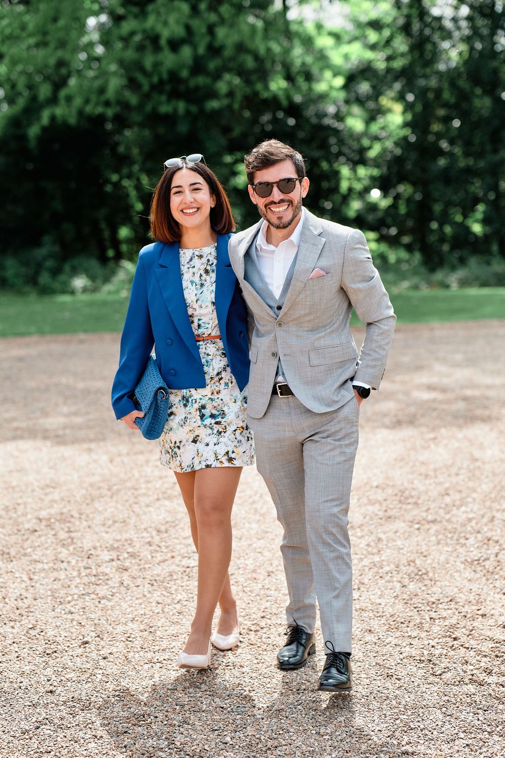 A couple in blue and gray attire shares joyful moments on a gravel path in an outdoor setting with lush greenery.