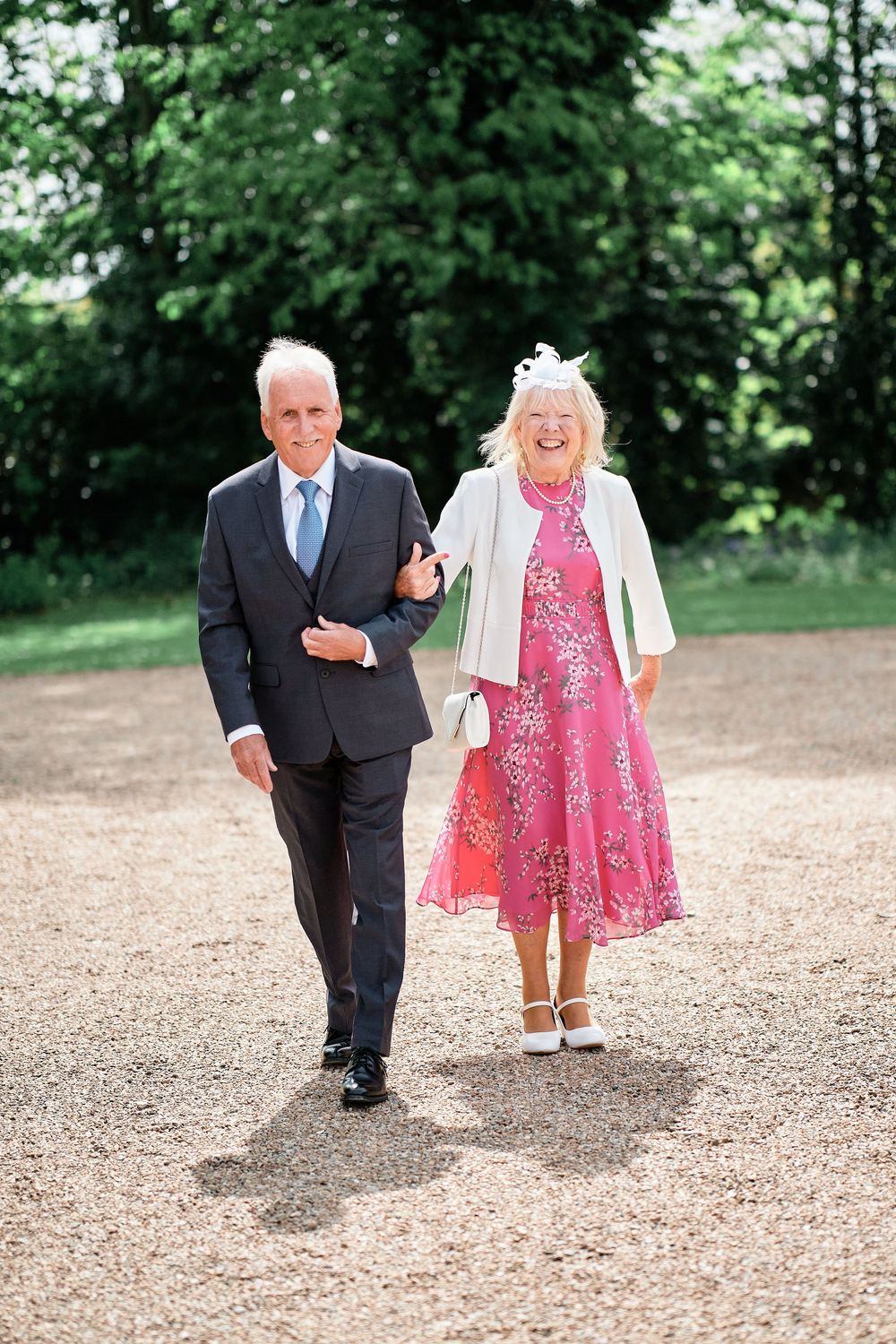 A couple walking arm in arm on a gravel path, the person on the right wearing a bright pink floral dress and white jacket.