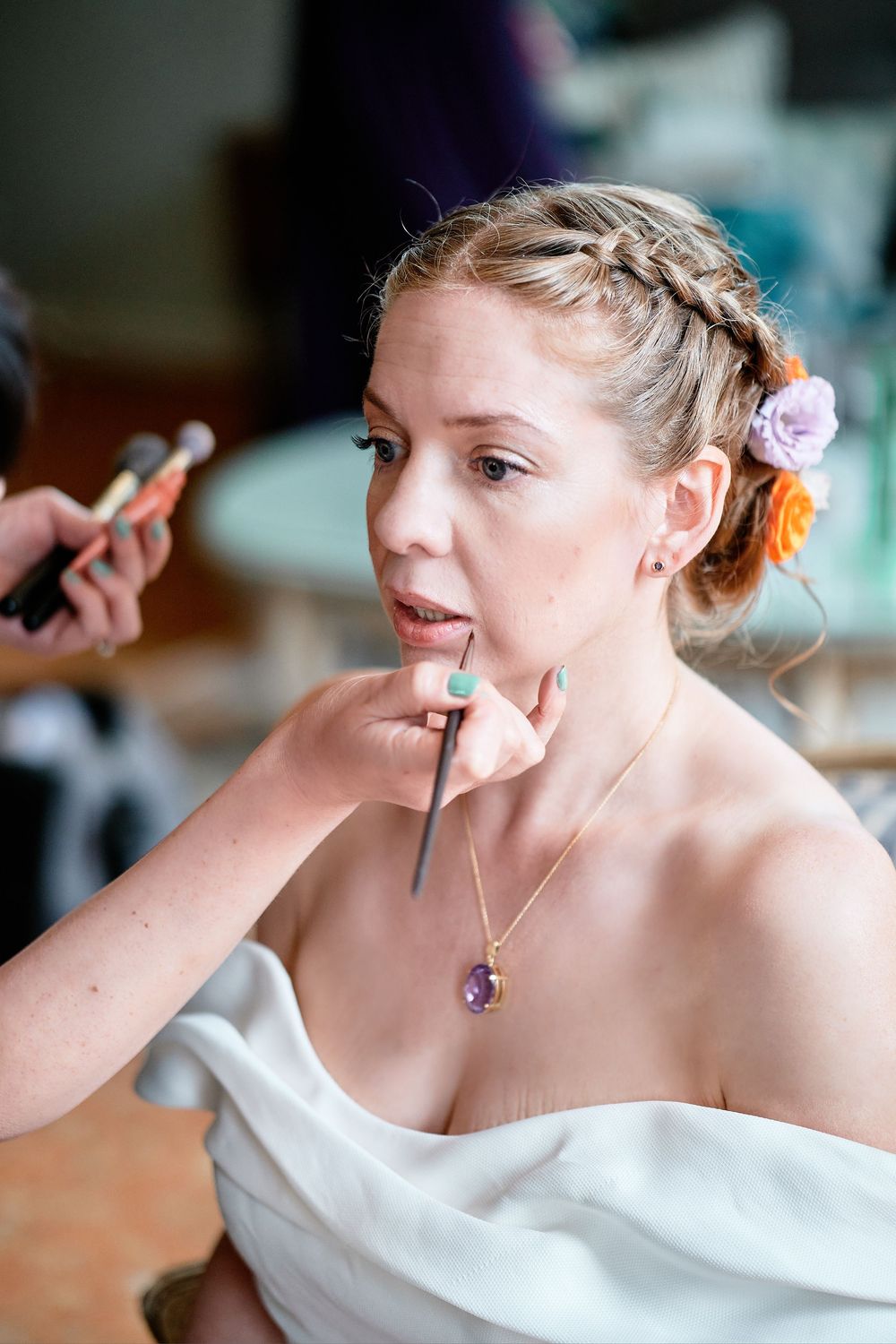 Makeup artist applies lipstick to bride in white dress with braided updo and pink flower during wedding preparation.