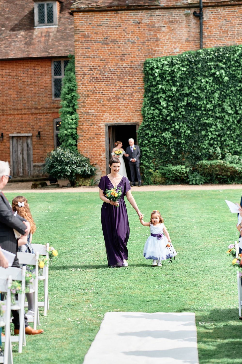 Bridesmaid in purple dress walks down outdoor wedding aisle with flower girl in white dress at brick venue with ivy.