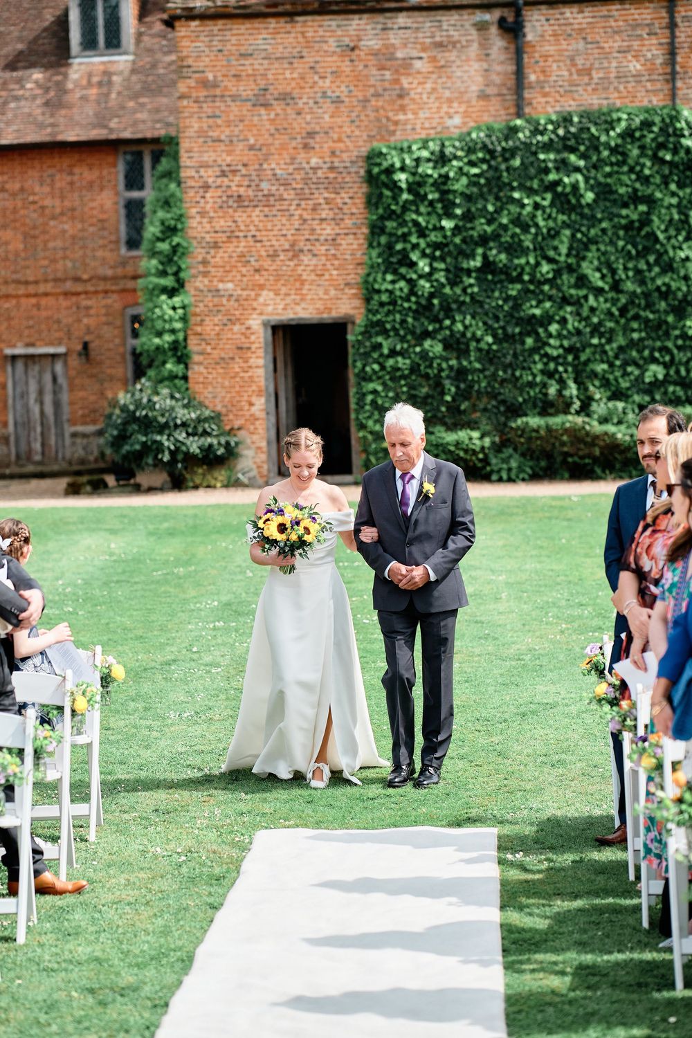A bride in white dress walks down an outdoor aisle with an escort on a lush lawn by a brick building.