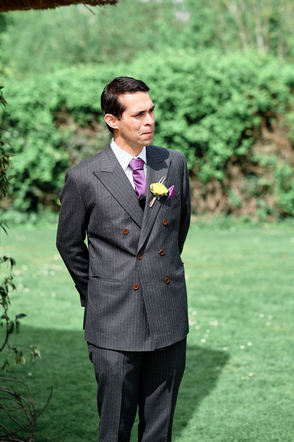 A wedding attendant in a pinstripe suit with purple accents stands on a grassy lawn during an outdoor ceremony.