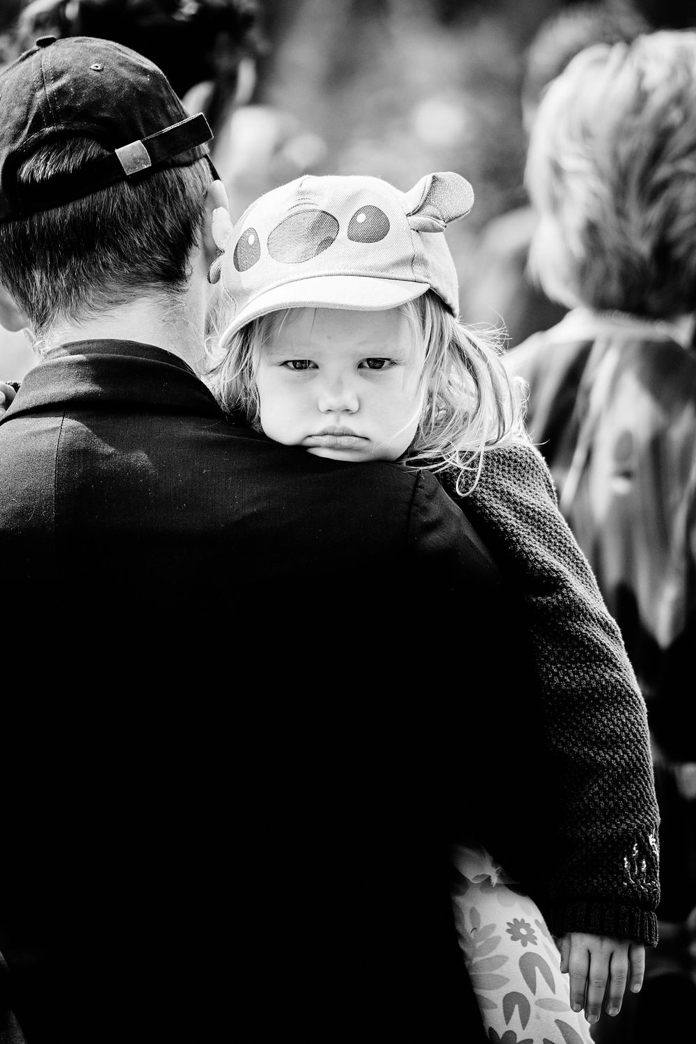 A black and white portrait of a child wearing an aviator-style cap while being carried on someone's shoulders.