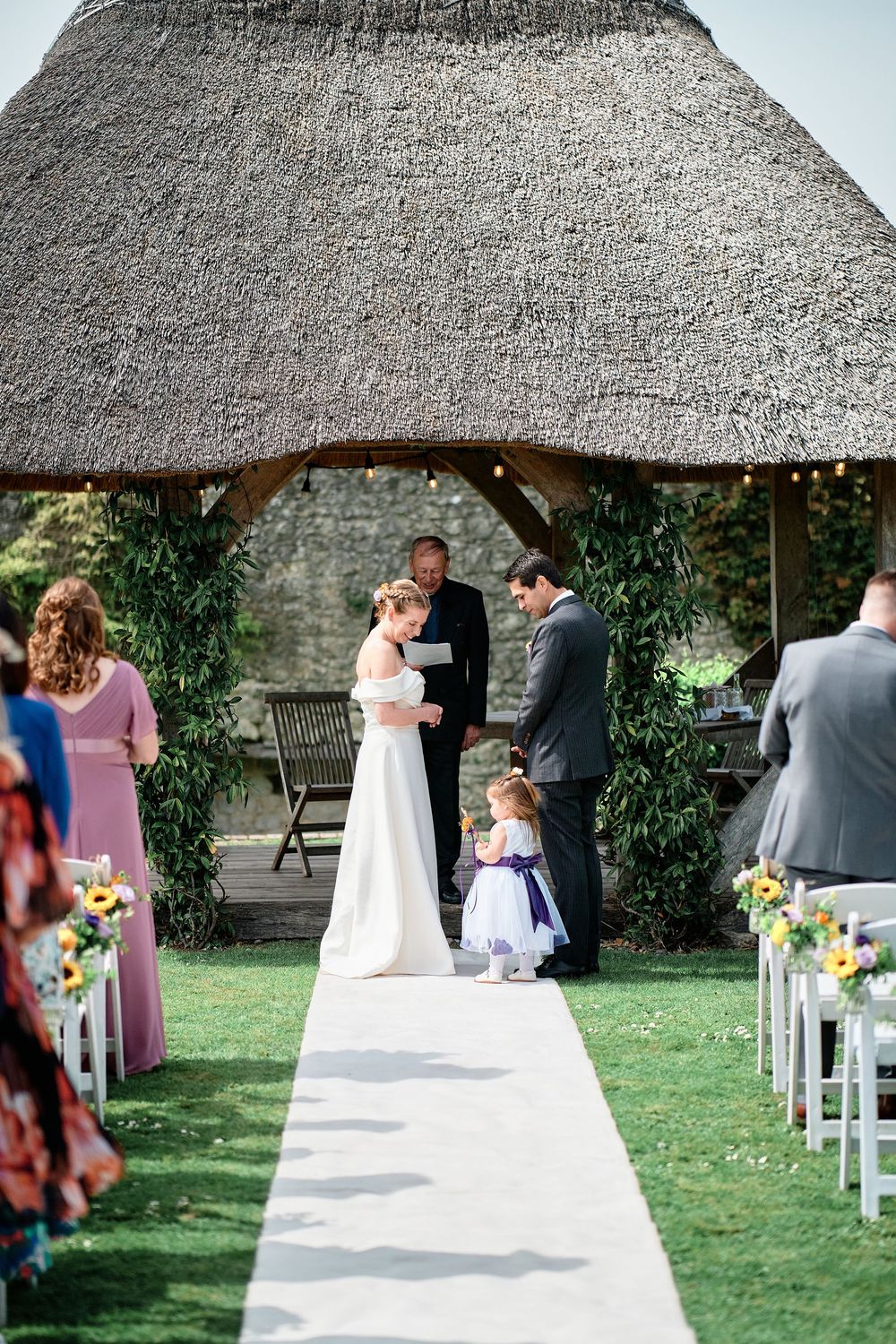 An outdoor wedding ceremony under a thatched gazebo with floral decorations and white chairs on a garden lawn.