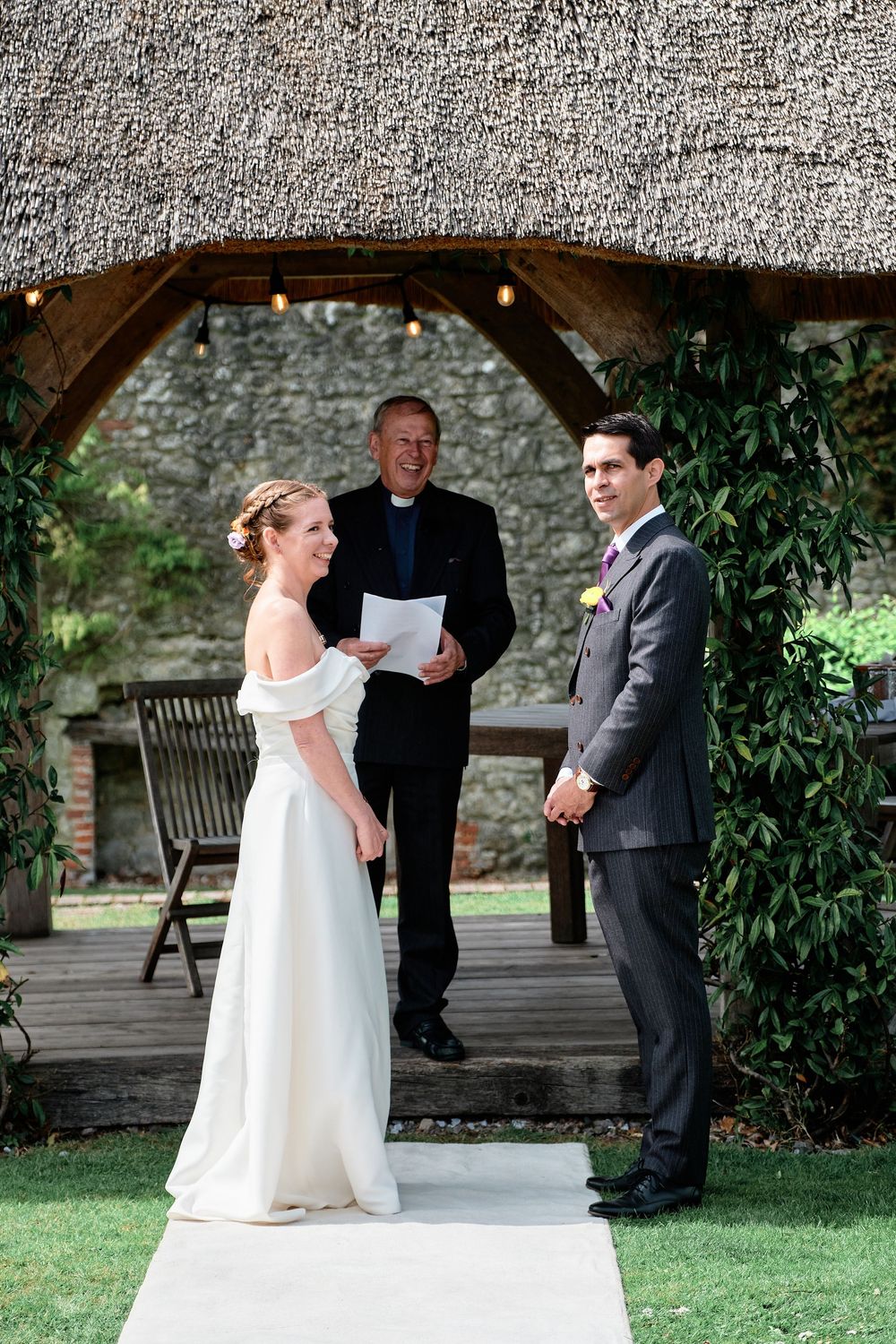 A couple stands with officiant under a rustic wooden archway during outdoor wedding ceremony.