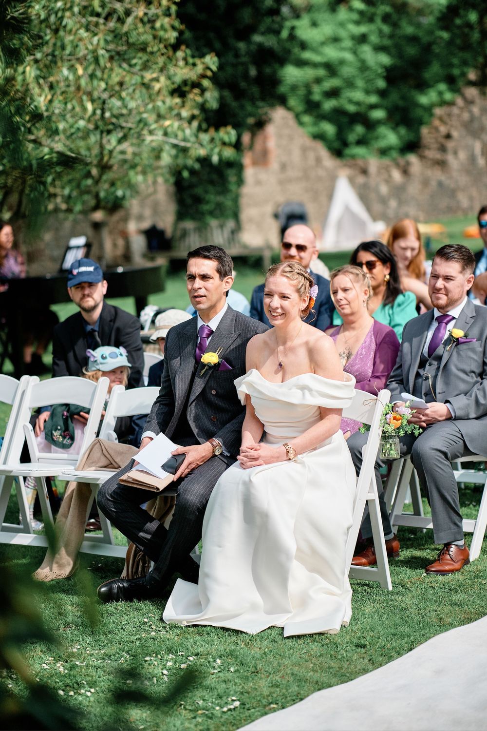 Guests seated at an outdoor wedding ceremony on white folding chairs watch joyfully during a sunny garden celebration.