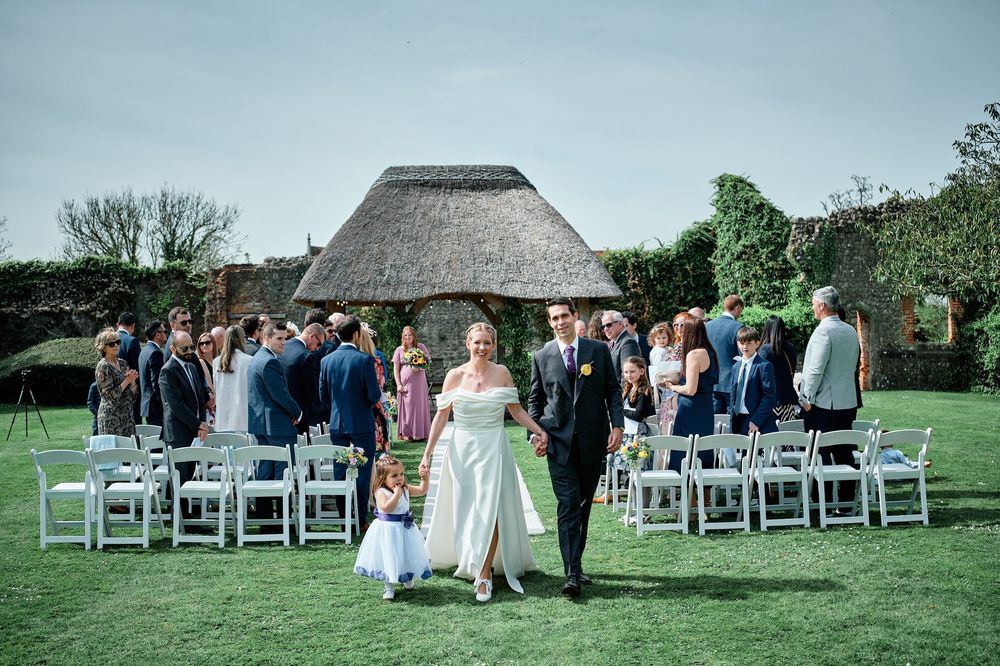 Wedding ceremony takes place on a lush green lawn with a rustic thatched-roof building in the background.