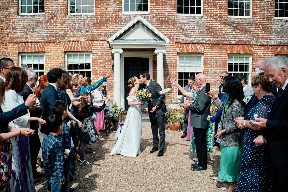 Wedding guests holding bubble wands form a celebratory exit line outside a historic brick mansion during a ceremony.