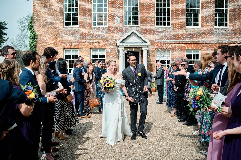 Wedding guests throw flower petals and confetti at a newlywed couple exiting a historic brick building.