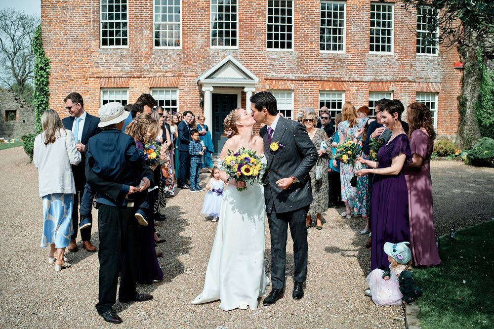 Wedding celebration outside a historic brick building with guests gathering and flowers on a sunny day.