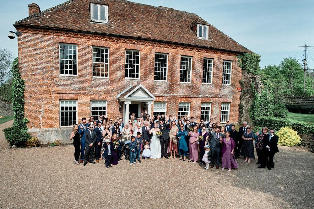 Large wedding party gathered outside a traditional Georgian brick house with symmetrical windows for a group photograph.