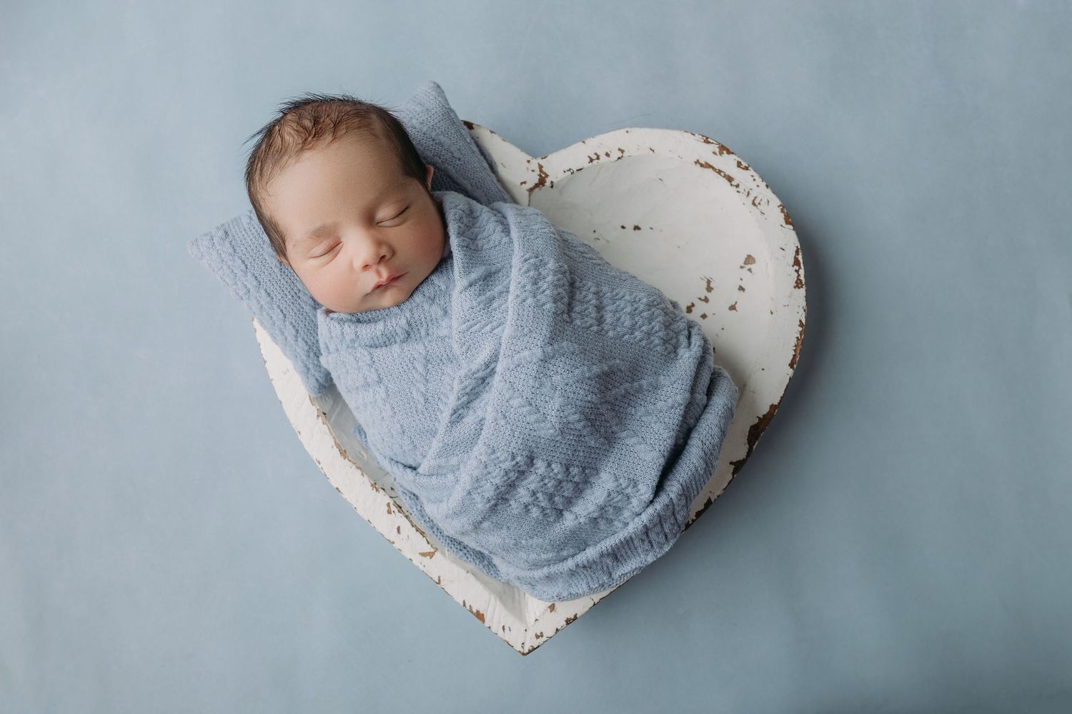 Sleeping baby wrapped in soft blue swaddle blanket lies peacefully in heart-shaped cushion on blue backdrop during newborn twins photos