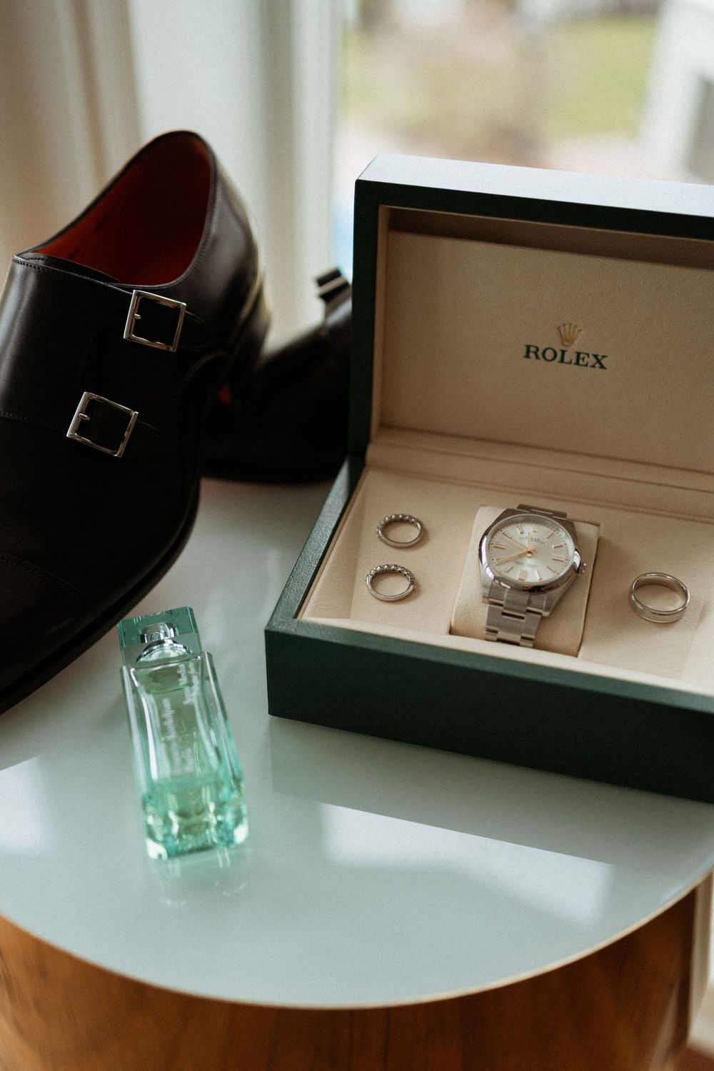 Luxury accessories including a black dress shoe, Rolex watch box with watch and rings, and green perfume bottle on white surface.