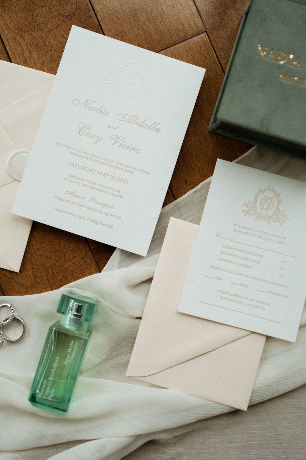 Elegant wedding invitation suite with gold lettering displayed alongside a green perfume bottle and white fabric.