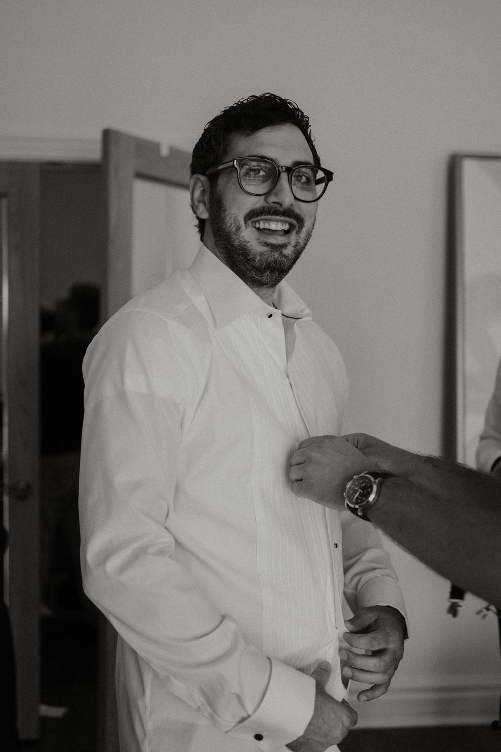 A groom wearing glasses and white dress shirt smiles while getting ready on his wedding day.