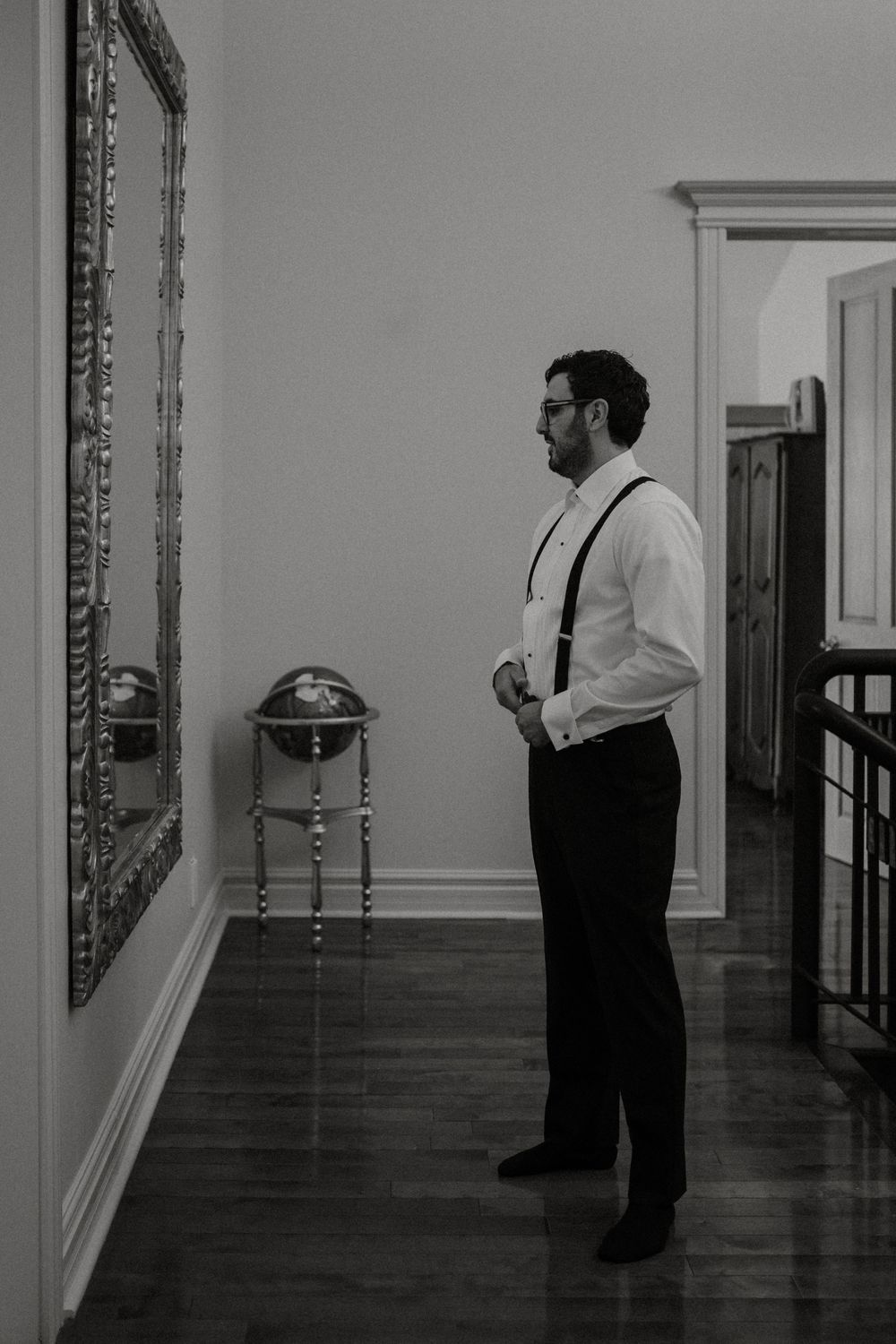 A well-dressed man in suspenders stands in an elegant hallway with hardwood floors and ornate mirror.