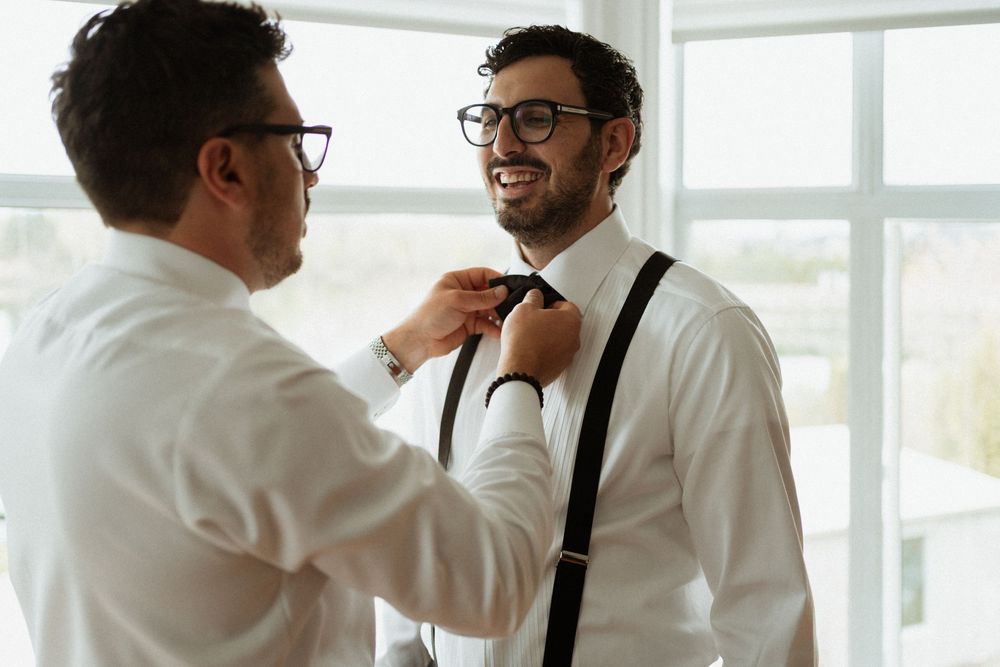 Two people dressed in white shirts and suspenders smile while adjusting a bow tie in a bright room.