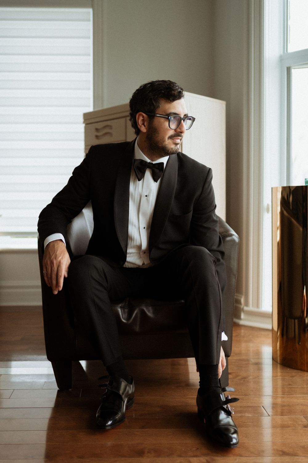 Man in classic black tuxedo and bow tie sits thoughtfully in a chair by a window.