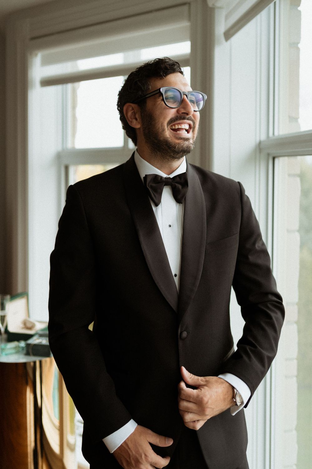 A groom in a black tuxedo and glasses laughs joyfully while standing by bright windows.
