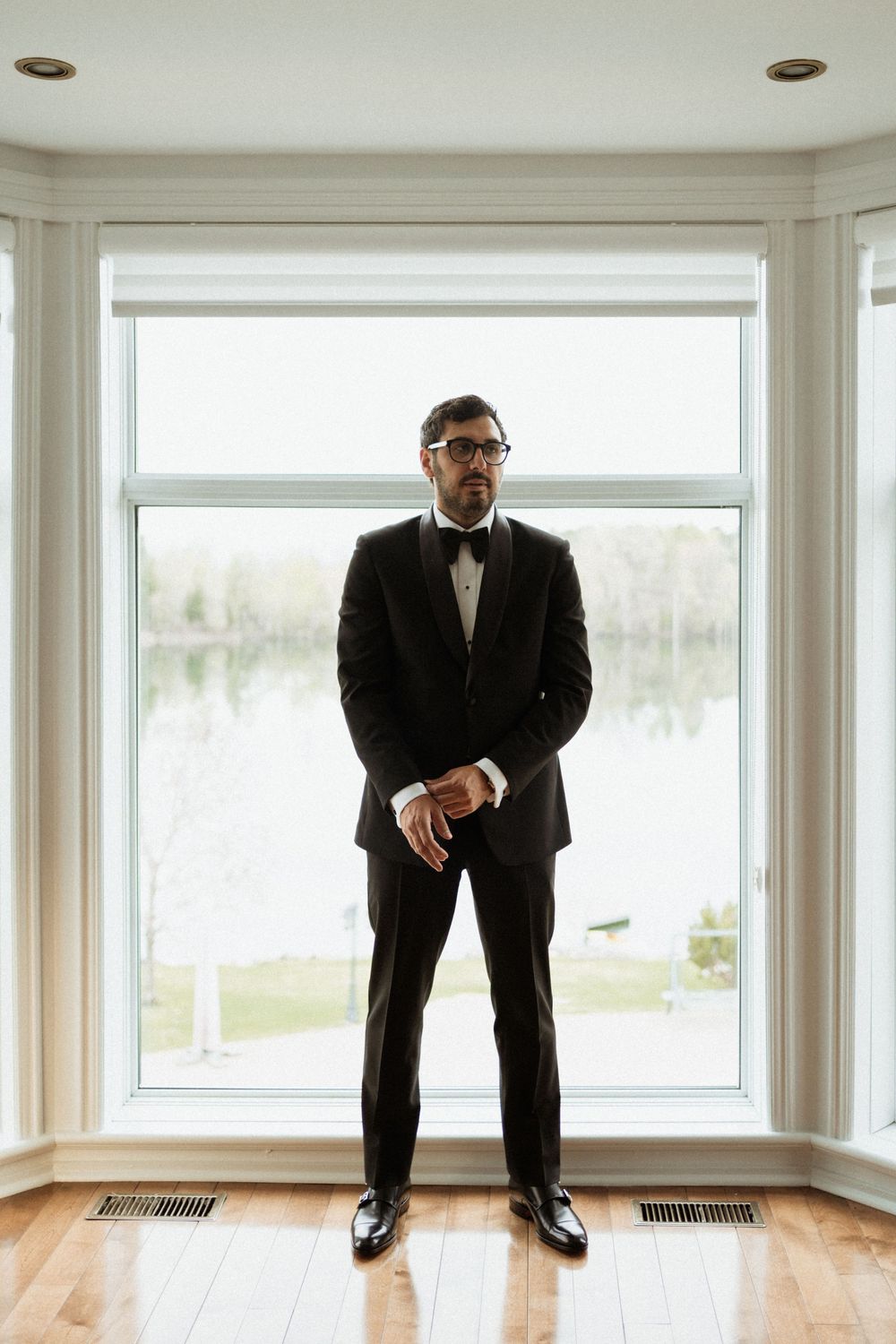 A groom in a classic black tuxedo and bow tie stands in front of a bright window in an elegant room.